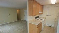 This single-floor space offers a functional and straightforward layout with an open connection between the kitchen and the adjacent living area. The kitchen is equipped with warm oak-toned cabinetry, including upper and lower storage cabinets that provide ample organization space. A white electric coil-top range and matching countertops in a light neutral finish give the kitchen a clean, practical look. The under-cabinet range hood adds a utilitarian touch above the cooking area. The kitchen transitions seamlessly into the carpeted living space, which features neutral beige wall-to-wall carpeting and matching painted walls that create a cohesive, understated atmosphere. A textured ceiling is present throughout, and the open sightline from the kitchen to the living area enhances the sense of connected space. Natural light appears to filter through the interior, contributing to a modest but comfortable ambiance. A doorway visible in the background suggests additional rooms beyond the main living space. The overall design is simple and functional, prioritizing practicality with neutral finishes suited to a variety of interior styles.