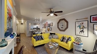 This single-floor Dallas residence offers an open-concept layout that connects the living area and kitchen in a cohesive, functional design. The living room serves as the focal point of the space, featuring crown molding, warm hardwood-style flooring, and a ceiling fan with integrated lighting that adds both comfort and practicality. A curated gallery wall with an assortment of framed artwork and a distinctive octagonal wood-framed mirror add character and visual depth to the neutral gray walls.

The kitchen is visible from the living area and features white cabinetry, a dark appliance suite, recessed lighting, and a breakfast bar that doubles as a casual dining spot. This open sightline enhances the sense of spaciousness throughout the unit. A dedicated dining area is positioned adjacent to the kitchen, making the layout ideal for entertaining.

Crown molding throughout adds a refined architectural touch, while the neutral color palette of the walls provides a versatile backdrop. Natural light filters in through windows near the entryway, brightening the interior. Overall, the space balances a modern, well-appointed design with a welcoming and lively atmosphere.