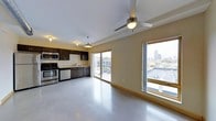 This single-floor unit features an open-plan layout that seamlessly integrates the kitchen and living area into one cohesive space. The kitchen is outfitted with dark espresso cabinetry, stainless steel appliances including a range and over-the-range microwave, and a streamlined countertop with a single-basin sink. Pendant lighting and flush-mounted ceiling fixtures illuminate the kitchen zone, while a ceiling fan with a light kit serves the broader living area, enhancing airflow and comfort throughout.

The living space is defined by polished concrete flooring that extends across the entire unit, lending an industrial yet modern aesthetic. Large windows, including a floor-to-ceiling sliding glass door, flood the interior with natural light and provide direct access to an outdoor terrace or balcony. The oversized windows frame elevated views of the surrounding cityscape and neighboring rooftops, offering a strong urban connection from within the unit. Neutral wall tones complement the overall palette, keeping the space bright and versatile. The open layout and generous fenestration give the unit a spacious, airy character well-suited to modern urban living.