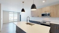 Located on North Arthur Ashe Boulevard in Richmond, Virginia, this thoughtfully designed 2-bedroom, 1-bathroom residence offers a cohesive modern aesthetic throughout its single floor. The open-concept kitchen and living area serves as the heart of the home, anchored by a large center island with a crisp white quartz countertop, an undermount sink, and a matte black spring-neck faucet. Bold navy-blue lower cabinetry with gold hardware contrasts elegantly with light wood upper cabinets, while a classic white subway tile backsplash and stainless steel appliances complete the chef-inspired space. Sleek pendant lights in black and brass hang above the island, adding a refined touch. Light hardwood flooring flows throughout the main living areas, enhancing the bright, airy ambiance. The primary bedroom offers generous proportions, large windows with outdoor views, and light hardwood flooring, while the second bedroom features plush carpeting, a ceiling fan, and similarly expansive windows that flood the room with natural light. A dedicated closet provides added storage. The full bathroom is finished with a navy vanity, gold hardware, white quartz countertop, a framed mirror, and a tub-shower combination with a rain showerhead — all tied together with consistent matte black accents.