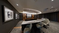 This upscale private screening room delivers a true cinema experience within a refined, well-appointed setting. The space is anchored by a large projection screen set against a curved wall of warm walnut wood paneling, flanked by decorative backlit panels and wall sconces that emit a soft, ambient glow. Floor-to-ceiling dark curtains line the walls, enhancing acoustic performance and contributing to the immersive, theater-like atmosphere.

Seating is arranged in two curved, tiered rows oriented toward the screen. The front row features a continuous curved bench with an integrated stone-topped counter, ideal for drinks or personal items, while the rear row offers generously sized, individual cognac leather armchairs with wooden frames — combining luxury with comfort. Step lighting along the tiered aisles ensures safe navigation in low-light conditions.

Overhead, a dramatic curved coffered ceiling with layered cove lighting serves as a striking architectural centerpiece, adding depth and elegance to the room. Recessed ceiling lights provide supplemental illumination. Textured charcoal carpeting grounds the space, and curated framed artwork adorns the dark accent walls, lending a gallery-like quality. A rear service counter with a decorative tile backsplash completes the room, making this an exceptional venue for private screenings and entertainment.