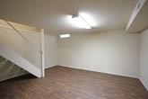 This finished basement presents a clean, open layout designed for flexible use. The flooring throughout consists of dark wood-look laminate planks that add warmth and a cohesive aesthetic to the below-grade space. The textured, popcorn-style ceilings are fitted with fluorescent tube lighting fixtures that provide bright, even illumination throughout the area.

A white-painted staircase with a railing serves as the primary access point, connecting this level to the floor above. The stairs feature carpet treads, contrasting with the smooth laminate flooring of the main space. Beneath and around the staircase, the open floor plan allows for a variety of configurations.

The walls are finished in a light, neutral tone with a subtle texture, and exposed structural beams are integrated cleanly into the ceiling design. HVAC vents are visible along the ceiling, indicating climate control throughout the space. A small egress window near the top of one wall allows a limited amount of natural light to filter in.

A separate door located at the base of the staircase provides access to an additional room or utility area, extending the functional footprint of the basement. Electrical outlets are distributed along the walls. Overall, the space offers a practical and well-finished environment suitable for storage, recreation, or additional living use.
