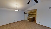 This single-floor residence offers a practical and comfortable layout centered around an open-concept living and kitchen area. The living space features neutral carpet flooring, clean white walls, and vaulted ceilings accented by a ceiling fan and a stylish pendant light fixture, contributing to an airy, spacious feel. The kitchen is seamlessly connected to the living area, creating an open and sociable floor plan. It is equipped with a full-size refrigerator, a range, and ample cabinetry finished in a warm, dark-trimmed cream tone. A central kitchen island provides additional counter space and serves as a natural divider between the cooking and living zones. Recessed and flush-mount ceiling lighting illuminates the kitchen effectively. The tile flooring in the kitchen contrasts with the carpeted living space, clearly delineating the two functional areas. The overall ambiance of the space is clean, understated, and move-in ready, with a neutral color palette throughout that allows for easy personalization. The layout is well-suited for everyday living, with the kitchen and living areas working together to support both casual dining and relaxed entertaining.