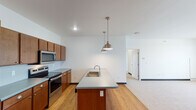 This well-designed single-floor unit in Fargo, North Dakota offers a thoughtfully laid-out living space with modern finishes throughout. The open-concept kitchen serves as the heart of the home, featuring rich warm-toned cabinetry, sleek light-gray countertops, and a full suite of stainless steel appliances including a refrigerator, electric range, and built-in microwave. A generously sized kitchen island with an integrated sink anchors the space, illuminated by stylish brushed nickel pendant lights that add a contemporary touch. Light hardwood flooring flows throughout the kitchen and main living areas, creating a warm and cohesive aesthetic.

The bedrooms are carpeted in neutral tones and bathed in natural light through windows fitted with horizontal blinds, offering views of the surrounding neighborhood. The rooms provide ample space for furnishings and personal use.

The bathroom is notably spacious, featuring dual sinks set into a continuous countertop with warm wood cabinetry below, a large mirror, and tile flooring. Tall storage cabinetry provides generous organizational space. The clean, neutral color palette of white walls and dark baseboards runs consistently throughout the unit, contributing to a bright, modern, and move-in-ready atmosphere.