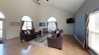 This single-floor space makes a strong first impression with its dramatic vaulted ceilings and open-concept layout that seamlessly connects multiple functional areas. The main living area is a standout, boasting soaring ceiling heights accentuated by a trio of tall arched windows that flood the room with abundant natural light. Comfortable seating arrangements, including plush sofas and patterned accent chairs, are thoughtfully arranged around a glass-top coffee table, while a wall-mounted television and track lighting add modern functionality. A secondary seating nook near the entryway features leather armchairs, a textured area rug, and additional high ceilings, reinforcing the airy atmosphere throughout. The dining area offers a sleek, industrial-style metal table and matching chairs set against a cool gray accent wall adorned with a coordinating triptych of abstract artwork. The kitchen is well-appointed with warm honey-toned wood cabinetry, granite-look countertops, a classic white subway tile backsplash, and black appliances including a refrigerator, range, and built-in microwave. Polished concrete-style flooring runs throughout the entire space, tying each area together with a cohesive, contemporary aesthetic.