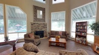 This single-story residence centers around a striking great room that immediately captures attention with its soaring two-story ceilings, creating an expansive and airy atmosphere. Large windows line the walls, flooding the space with natural light and offering pleasant views of the surrounding greenery and outdoor pool area. A ceiling fan overhead ensures year-round comfort in this generous living space.

The focal point of the great room is a beautifully crafted stacked stone corner fireplace, lending warmth and a rustic charm to the otherwise neutral, contemporary interior. Above the fireplace mantle, an ornate decorative mirror adds an artistic touch to the space. The room is finished with neutral carpeting and warm beige walls, providing a versatile backdrop for the comfortable, traditional furnishings arranged throughout.

A wooden coffee table anchors the seating area, while a built-in bookcase along one wall adds both function and character. The combination of the stone fireplace, dramatic ceiling height, and abundant natural light gives this home a welcoming and refined ambiance, making the great room a true centerpiece of everyday living and entertaining.