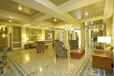 Located at 145 Granby Street in Norfolk, Virginia, this single-floor space presents an elegantly designed lobby and common area that balances classical architectural detail with contemporary furnishings. The layout is expansive, organized along a central corridor framed by prominent white columns that draw the eye through the length of the space, creating a grand sense of depth and proportion.

The polished terrazzo flooring is one of the most striking permanent features, its speckled surface reflecting the warm glow of multiple chandeliers suspended from coffered ceilings throughout. Decorative inlay borders along the floor accentuate the formal layout and add visual interest underfoot. The coffered ceiling design, complemented by recessed lighting and classical crown molding, reinforces the upscale architectural character of the space.

Distinct zones are established throughout, including a lounge area furnished with plush upholstered armchairs arranged around low tables, an office or concierge area featuring a walnut credenza and a large ornate mirror, and a mail room alcove with built-in mailboxes. A grand piano is prominently positioned near the street-facing entrance, adding a cultural and performative element. Dark wood flooring transitions the space into secondary corridors, where a staircase with metal railings provides additional vertical access. Curated artwork is displayed throughout, contributing to the polished, hospitality-inspired ambiance.