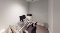 This study room is a compact, single-level space designed to support focused individual work and computing tasks. The room features neutral light grey walls, carpeted flooring, and clean white baseboards that contribute to a simple, functional aesthetic. A glass-and-metal desk serves as the central workstation, equipped with a desktop monitor, keyboard, and mouse, providing a dedicated computing setup for users. A printer is positioned on a separate white shelf unit adjacent to the desk, offering convenient document handling capabilities. An ergonomic office chair accompanies the workstation, ensuring comfortable use during extended sessions. A small waste bin is also present, adding to the room's practical furnishings. The space is monitored by a ceiling-mounted security camera, and posted signage reinforces no-smoking policies and notifies users of active surveillance. Narrow horizontal windows positioned high on the wall allow a modest amount of natural light to filter in while maintaining privacy. Instructional notices are displayed on the wall, providing users with guidance on operating the equipment and connecting to the local network. Overall, the room offers a straightforward, well-equipped environment suited for individual study or computer-based tasks.