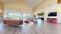 This expansive single-story community clubhouse offers a thoughtfully designed layout across a generous floor plan. The grand entrance welcomes visitors through soaring floor-to-ceiling glass doors, opening into a bright, airy foyer complete with a reception desk and large windows that invite abundant natural light. The centerpiece of the facility is a dramatic great room with soaring vaulted ceilings, warm wood-look flooring, and an impressive wall of large-pane windows that frame outdoor views and flood the space with daylight. Comfortable seating arrangements and a chandelier pendant light create an inviting, relaxed atmosphere. Adjacent to the living area, a combined dining and living space features high-top seating and direct access to an outdoor terrace through expansive sliding glass doors. The well-appointed kitchen is outfitted with dark wood cabinetry, stainless steel appliances, granite countertops, and a large window overlooking the exterior. A dedicated game room offers a classic billiard table beneath a coffered ceiling with dark wood beams. The media room, also featuring a coffered ceiling, provides a comfortable theater-style seating area. Completing the amenities, a fully equipped exercise room features cardio machines, strength equipment, and a vibrant accent wall, ensuring a comprehensive fitness experience throughout this versatile community space.