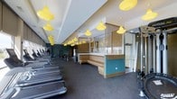 This single-floor fitness center combines functionality with a thoughtfully designed aesthetic. The space is anchored by a striking array of bold yellow pendant lights that hang from a recessed ceiling, adding a vibrant and energetic ambiance throughout. The flooring is finished in durable dark rubber matting, ideal for high-traffic workout areas.

Along one side of the space, a row of treadmills and cardio machines is neatly arranged, offering ample equipment for aerobic exercise. Toward the rear, strength training machines and additional fitness equipment are visible, providing a comprehensive workout environment. The dark teal accent walls in the training area create a focused, dynamic atmosphere.

A distinctive lounge or reception area is centrally positioned, featuring wraparound upholstered booth seating in warm tones, framed by a teal base and accented with decorative pendant lighting including a glamorous globe chandelier. Woven bamboo-style blinds and glass partitions define this area while maintaining an open feel.

Natural light filters in through large windows, brightening the space and complementing the warm artificial lighting. The overall design balances an inviting social zone with a fully functional workout facility, making this fitness center both practical and visually engaging.