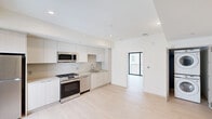 This well-appointed one-bedroom, one-bathroom residence offers a thoughtfully designed layout across a single floor. The open-plan living and kitchen area serves as the heart of the unit, featuring light-toned hardwood flooring that extends throughout and recessed lighting that keeps the space bright and airy. The modern kitchen is equipped with a full suite of stainless steel appliances, including a gas range, over-the-range microwave, dishwasher, and refrigerator. Flat-panel cabinetry in a soft white finish reaches toward the ceiling, maximizing storage, while a single-basin sink and clean countertops complete the functional workspace. A large window and sliding glass door flood the living area with natural light and provide direct access to a private balcony enclosed by a sleek metal railing. The bedroom offers a comfortable footprint with the same light hardwood flooring and a sliding glass door leading to the balcony, enhancing the sense of openness. The bathroom is finished with large-format neutral tile, a glass-enclosed walk-in shower with a built-in niche, matte black hardware accents, an undermount sink set in a white vanity, and a wide mirror. An in-unit stacked washer and dryer is also included, adding everyday convenience to this clean, contemporary residence.