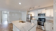 Located at 3801 Banyan Grove Ln in Virginia Beach, VA, this 3-bedroom, single-floor unit offers a clean, modern design with a neutral color palette and thoughtful finishes throughout. The open-concept kitchen is a standout feature, showcasing white shaker-style cabinetry, granite countertops with subtle veining, a large center island with an undermount double sink and gold-tone faucet, and a full suite of stainless steel appliances including a gas range, built-in microwave, and refrigerator. A classic tile backsplash and track lighting complete the kitchen's polished look. The living and dining areas flow naturally from the kitchen, featuring warm wood-style flooring, crown molding details, and recessed lighting that keeps the space bright and welcoming. A dedicated entryway provides a clean transition into the main living space. The bedrooms are carpeted for comfort and receive natural light through windows with blinds, and closet access is provided in each room. The bathrooms are well-appointed with granite vanity tops, white cabinetry with brushed nickel hardware, soaking tub and shower combinations with tiled surrounds, and matching wood-style flooring. The overall ambiance is fresh, clean, and move-in ready.
