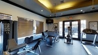 This single-floor fitness center is thoughtfully designed to offer a complete workout experience in a comfortable, well-appointed environment. The space features a distinctive tray ceiling with warm, recessed lighting and a ceiling fan, creating an inviting atmosphere that encourages activity. The walls are finished in a neutral tone, accented by bold dark trim that adds a modern, polished character to the room.

The facility is equipped with a selection of cardio machines, including elliptical trainers and a stationary bike, as well as strength training equipment such as a leg press and weight bench, catering to a variety of fitness routines. A wall-mounted flat-screen television provides entertainment during workouts, while durable rubberized flooring ensures safety and comfort underfoot.

Natural light flows in generously through large windows fitted with horizontal blinds, offering control over brightness and privacy. A set of glass-paned double doors opens directly to an outdoor area, allowing for ventilation and a connection to the exterior environment. The overall ambiance strikes a balance between functional utility and contemporary design, making this fitness center a practical and appealing space for daily exercise.