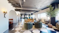 Located at 1020 N Tyndall Ave in Tucson, AZ, this thoughtfully designed communal space occupies a generous single floor and masterfully combines industrial aesthetics with vibrant, eclectic interiors. Bold black-and-white and blue-and-white checkered tile flooring anchors the main open area, setting a striking visual tone throughout. Exposed concrete ceilings with visible ductwork and conduit lend an urban, loft-like character, while large floor-to-ceiling windows flood the space with abundant natural light and frame panoramic views of the Tucson skyline and surrounding landscape.

The main lounge area is furnished with plush velvet barrel chairs, a tufted leather sofa, and layered area rugs, creating multiple intimate seating arrangements. Arched architectural details frame a cozy built-in banquette with a round bistro table, adding a refined, boutique touch. A bar-height counter with stools and a gallery wall of framed artwork anchors a casual café-style zone, while a dedicated dining area features bistro tables, bentwood chairs, and potted olive trees for a lively, café-inspired atmosphere.

A separate, richly appointed room features deep burgundy walls and ceiling, herringbone wood flooring, a dramatic floor-to-ceiling bookshelf mural, a Chesterfield sofa, and a conference table — ideal as a private study or meeting room. Multiple hallways connect the distinct zones seamlessly throughout the floor.
