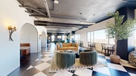 Located at 1020 N Tyndall Ave in Tucson, AZ, this thoughtfully designed communal space occupies a generous single floor and masterfully combines industrial aesthetics with vibrant, eclectic interiors. Bold black-and-white and blue-and-white checkered tile flooring anchors the main open area, setting a striking visual tone throughout. Exposed concrete ceilings with visible ductwork and conduit lend an urban, loft-like character, while large floor-to-ceiling windows flood the space with abundant natural light and frame panoramic views of the Tucson skyline and surrounding landscape.

The main lounge area is furnished with plush velvet barrel chairs, a tufted leather sofa, and layered area rugs, creating multiple intimate seating arrangements. Arched architectural details frame a cozy built-in banquette with a round bistro table, adding a refined, boutique touch. A bar-height counter with stools and a gallery wall of framed artwork anchors a casual café-style zone, while a dedicated dining area features bistro tables, bentwood chairs, and potted olive trees for a lively, café-inspired atmosphere.

A separate, richly appointed room features deep burgundy walls and ceiling, herringbone wood flooring, a dramatic floor-to-ceiling bookshelf mural, a Chesterfield sofa, and a conference table — ideal as a private study or meeting room. Multiple hallways connect the distinct zones seamlessly throughout the floor.