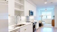 This thoughtfully laid-out studio residence offers a clean, modern aesthetic throughout its single floor. The open-concept kitchen and living area form the heart of the unit, featuring a galley-style kitchen outfitted with crisp white cabinetry accented by gold hardware, a subway tile backsplash, stainless steel appliances including a range, microwave, and refrigerator, and a deep undermount sink with a professional-style faucet. Light wood-tone flooring runs throughout the main living areas, enhancing the bright, airy feel. The living space is framed by a large window that invites natural light and provides outdoor views, complemented by a ceiling fan overhead. The bedroom is a comfortable and private retreat, also featuring a ceiling fan and natural light. Two closets provide generous storage, including a spacious walk-in closet with built-in shelving and dual hanging rods, finished with plush carpet underfoot. The bathroom offers a clean, elongated layout with large-format tile flooring, a white vanity with gold hardware, a backlit mirror, and a full bathtub with shower. The entry foyer includes built-in open shelving for added convenience. Recessed lighting and an in-unit washer and dryer round out the practical amenities of this well-appointed residence.