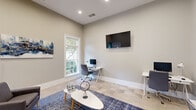 Located at 7305 Village Center Blvd in Fairburn, GA, this single-floor space is a well-appointed multipurpose room that blends workspace functionality with comfortable lounge elements. The room features soft, neutral gray walls complemented by light-toned tile flooring that runs throughout, creating a clean and cohesive aesthetic.

Natural light enters generously through large windows, offering views of surrounding greenery and lending the room an open, airy feel despite its intimate footprint. Recessed ceiling lighting supplements the natural light, ensuring the space remains bright and welcoming throughout the day.

The workspace area includes multiple white desks paired with ergonomic gray chairs and desktop computers, making it well-suited for focused work or collaboration. A wall-mounted flat-screen television is prominently positioned, adding a media and presentation capability to the room.

A central lounge area anchors the space with a bold blue patterned area rug, a modern oval coffee table, and upholstered gray accent chairs, creating a casual gathering zone. Decorative artwork, including a vibrant abstract canvas and a striking blue-and-gold world map triptych, adds personality and visual interest to the walls.

A hallway leads to an accessible restroom, and a glass-panel exterior door provides direct access to the outdoors. This thoughtfully designed space is versatile, serving equally well as a business center, coworking lounge, or community meeting room.