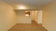 This single-floor residential space offers a practical and straightforward layout designed for comfortable everyday living. The living area is finished with wall-to-wall carpet, providing a warm and soft underfoot feel, and is illuminated by a flush-mount ceiling light. A standout architectural detail is the pass-through window cut into the dividing wall between the living area and the kitchen, allowing for easy communication and light sharing between the two spaces. The kitchen features dark wood cabinetry and is partially visible through this opening, hinting at a functional cooking space beyond. Transitioning from the carpeted living area, the flooring shifts to hardwood-style planks in the hallway, creating a clear visual distinction between zones. The hallway leads to additional rooms and features a louvered closet door, offering convenient storage solutions. The walls throughout are finished in a neutral, light tone that contributes to an open and airy ambiance, while the textured ceiling adds a subtle layer of detail. Overall, the space presents a clean, no-frills interior with practical design choices suited for daily residential use.