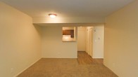 This single-floor residential space offers a practical and straightforward layout designed for comfortable everyday living. The living area is finished with wall-to-wall carpet, providing a warm and soft underfoot feel, and is illuminated by a flush-mount ceiling light. A standout architectural detail is the pass-through window cut into the dividing wall between the living area and the kitchen, allowing for easy communication and light sharing between the two spaces. The kitchen features dark wood cabinetry and is partially visible through this opening, hinting at a functional cooking space beyond. Transitioning from the carpeted living area, the flooring shifts to hardwood-style planks in the hallway, creating a clear visual distinction between zones. The hallway leads to additional rooms and features a louvered closet door, offering convenient storage solutions. The walls throughout are finished in a neutral, light tone that contributes to an open and airy ambiance, while the textured ceiling adds a subtle layer of detail. Overall, the space presents a clean, no-frills interior with practical design choices suited for daily residential use.