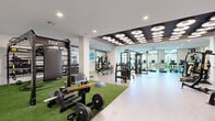 Located at 5905 Caravan Ct in Orlando, FL, this professionally appointed fitness center spans a generously sized single floor, offering a well-organized and fully equipped exercise environment. The space is divided into distinct training zones, each designed to accommodate a wide variety of workout styles and fitness goals.

The main floor features light wood-toned flooring throughout, accented by zones of green turf and yellow-green rubberized flooring that visually separate functional areas. A standout architectural feature is the bold, dark ceiling panel adorned with circular pendant light clusters, providing dramatic and focused illumination over the strength training zone. Recessed lighting throughout the remainder of the space ensures consistent, bright conditions.

The strength training area is outfitted with a comprehensive selection of selectorized machines, cable systems, TRX suspension trainers, free weights, and barbells. A dedicated turf section supports functional and dynamic training exercises. The cardio zone is lined with treadmills, stationary bikes—including Peloton-style cycles—and other cardio equipment, positioned near floor-to-ceiling windows that offer views of lush outdoor greenery and palm trees, filling the space with natural light.

A separate glass-enclosed studio room provides a private space for group classes or personal training. Large mirrors, wall-mounted televisions, and curated artwork complete the modern, motivating atmosphere of this exceptional fitness facility.