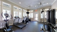 This fitness room offers a dedicated space for exercise, thoughtfully designed with both functionality and comfort in mind. The room is finished with warm beige walls, white crown molding, and recessed lighting, giving it a clean, polished appearance. Two ceiling fans ensure consistent air circulation throughout the space during workouts.

Natural light fills the room generously through multiple large windows positioned along the walls, offering views of surrounding greenery and creating an open, welcoming atmosphere. A glass-paneled door provides additional light and convenient access.

The fitness area is equipped with a treadmill, two stationary bikes, and a multi-function cable weight machine, catering to both cardiovascular and strength training needs. A wall-mounted flat-screen television is positioned centrally for entertainment during workouts. The floor is covered in durable rubber matting, providing safety, noise reduction, and cushioning underfoot.

An illuminated exit sign highlights the room's compliance with safety standards. Overall, the space delivers a practical and comfortable environment suited for a consistent fitness routine.