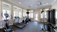 This fitness room offers a dedicated space for exercise, thoughtfully designed with both functionality and comfort in mind. The room is finished with warm beige walls, white crown molding, and recessed lighting, giving it a clean, polished appearance. Two ceiling fans ensure consistent air circulation throughout the space during workouts.

Natural light fills the room generously through multiple large windows positioned along the walls, offering views of surrounding greenery and creating an open, welcoming atmosphere. A glass-paneled door provides additional light and convenient access.

The fitness area is equipped with a treadmill, two stationary bikes, and a multi-function cable weight machine, catering to both cardiovascular and strength training needs. A wall-mounted flat-screen television is positioned centrally for entertainment during workouts. The floor is covered in durable rubber matting, providing safety, noise reduction, and cushioning underfoot.

An illuminated exit sign highlights the room's compliance with safety standards. Overall, the space delivers a practical and comfortable environment suited for a consistent fitness routine.