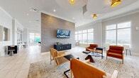 This expansive single-floor amenity space is thoughtfully designed to accommodate a variety of activities within a cohesive, contemporary aesthetic. The layout is divided into several functional zones that transition seamlessly from one to the next. The main lounge area serves as the centerpiece, featuring a textured tile accent wall with a wall-mounted television, a media console, and a curated arrangement of cognac leather and cane-back chairs set atop a patterned area rug. Overhead, sculptural tiered ceiling details and woven pendant light fixtures add architectural interest. Large windows throughout flood the space with natural light while framing panoramic city views. An entertainment zone houses a full-length shuffleboard table and a foosball table, complemented by additional woven pendant lighting and direct access to an outdoor area. A bar-height island with a dark stone surface and leather stools anchors a social gathering area equipped with a wet bar, beverage refrigerator, and wall-mounted display. Light-toned large-format tile flooring in a herringbone pattern unifies this section. The co-working area offers built-in workstations, ample desktop space, and comfortable seating, while a glass-walled private conference room provides a dedicated space for focused meetings. Botanical artwork and neutral-toned furnishings throughout contribute to a polished, welcoming atmosphere.