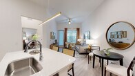 This thoughtfully designed 1-bedroom, 1-bathroom residence offers an efficient yet comfortable layout across a single floor. The open-concept kitchen and living area serves as the heart of the home, featuring soaring ceilings, light wood-style flooring, and a ceiling fan that enhances the sense of airiness. The kitchen is outfitted with dark shaker-style cabinetry, white quartz countertops, a hexagonal tile backsplash, stainless steel appliances including a refrigerator, range, and microwave, and a center island with seating for two—ideal for casual dining or entertaining. A gold linear pendant light adds a refined touch overhead. The living area is bathed in natural light from large windows dressed with elegant drapes, and a glass door provides access to an outdoor balcony. The bedroom offers a cozy retreat with plush carpeting, a bright window, and ample wall space. The full bathroom includes a dark-cabinet vanity with a white countertop, a large mirror, and a tub-shower combination. A hallway and dedicated closet round out the layout, providing practical storage solutions. Consistent white walls and gold accent details create a cohesive, modern aesthetic throughout.