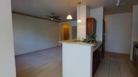 This single-story space showcases an open-concept design that seamlessly connects the kitchen and living area, creating a cohesive and functional environment. The kitchen is anchored by a peninsula with a light-toned countertop, offering ample workspace and a casual seating or serving area. Dark-stained wood cabinetry provides rich contrast against the neutral surroundings, while a built-in sink with a modern faucet adds practicality. Two pendant lights hang above the peninsula, casting warm, focused illumination over the work surface.

The adjoining living area features light-toned flooring that transitions smoothly from the tile in the kitchen, and a ceiling fan overhead ensures comfortable airflow throughout. Crown molding adds a refined architectural detail along the upper walls. A distinctive arched doorway frames the entry foyer in the background, lending a classic design element to the space. The front entry, visible through the arch, features a bold-colored door that provides a subtle focal point. The overall palette of warm neutrals and wood tones gives the space an inviting, well-maintained ambiance.