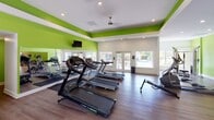 Located at 1916 Paces Landing Ave in Rock Hill, SC, this single-floor fitness facility is designed to support a comprehensive wellness experience. The main workout area is immediately identifiable by its bold lime-green accent walls paired with crisp white ceilings, creating an energetic and motivating atmosphere. Durable wood-look flooring runs throughout, offering both practicality and a polished aesthetic.

The primary exercise space is generously sized and well-lit, combining recessed ceiling lighting with abundant natural light from large windows and glass-paned double doors that offer views of the surrounding greenery. Full-length mirrors along one wall visually expand the space while aiding exercisers with form and technique. The room is equipped with multiple treadmills, elliptical machines, recumbent and upright bikes, a rowing machine, and a rack of dumbbells. Wall-mounted televisions provide entertainment during workouts, and a ceiling fan ensures comfortable airflow.

A secondary area connects seamlessly to the main floor, housing additional strength and cardio equipment with the same consistent design language of green walls and wood flooring. A separate, dedicated children's room features playful typographic wallpaper, colorful child-sized furniture, a striped area rug, and a window overlooking the outdoors—making the facility accommodating for families.