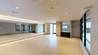 This fitness yoga studio is designed to support a focused and functional workout environment. The open floor plan provides ample space for group classes or individual practice, finished with smooth, light-toned flooring that is easy to clean and comfortable underfoot. A full-length mirrored wall along one side enhances the sense of space while allowing practitioners to monitor their form during sessions. A ballet barre runs along the mirrored wall, adding versatility for barre-based workouts and stretching routines. Recessed lighting and a softly illuminated ceiling cove provide even, adjustable illumination throughout the room, contributing to a calm and focused atmosphere. A wall-mounted flat-screen display is positioned at the front of the studio, useful for streaming guided classes or displaying instructor content. Built-in cubby-style storage beneath the screen offers organized space for yoga mats, blocks, and other fitness accessories. A window along the back wall allows natural light to enter, brightening the space and creating a welcoming ambiance. The neutral color palette and clean architectural lines give the studio a modern, professional feel suited for a variety of movement-based disciplines.