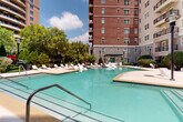 This outdoor amenity space is thoughtfully designed to serve as a central gathering hub within a multi-story residential complex. The architectural backdrop features a striking combination of classic red brick and natural stone facades rising several stories, with private balconies overlooking the courtyard below, creating an enclosed and intimate atmosphere.

The centerpiece of the space is a generously sized, freeform swimming pool with clear turquoise water, framed by a wide, smooth concrete sun deck. Rows of white lounge chairs are arranged along the pool's perimeter, providing ample space for sunbathing and relaxation. Entry points to the pool are equipped with stainless steel handrails for easy access.

Beyond the pool, the courtyard transitions into thoughtfully landscaped zones featuring raised planters with flowering shrubs, ornamental grasses, and mature trees that soften the hardscape. Modern angular outdoor furniture, including geometric chairs and clean-lined tables, is positioned throughout the patio areas, offering casual seating beneath open skies.

Classic black lamp posts punctuate the walkways, enhancing the space's traditional aesthetic while providing evening illumination. Decorative stone-bordered garden beds filled with mulch and seasonal plantings add color and texture throughout, contributing to a well-maintained, resort-inspired ambiance.
