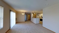 This single-floor space is designed around an open-concept layout that seamlessly connects the living area and kitchen, creating a bright and functional environment. The living area is finished with plush wall-to-wall carpeting in a neutral tone, contributing to a warm and comfortable atmosphere. A pendant light fixture hangs overhead, providing focused illumination, while a large window fitted with vertical blinds allows natural light to filter into the space.

The kitchen is positioned adjacent to the living area and is defined by a wraparound peninsula that serves as both a workspace and a natural divider between the two zones. White cabinetry lines the kitchen walls, complemented by a multi-bulb pendant light and a track lighting system above. Standard appliances, including a refrigerator, electric range, and microwave, are included. The kitchen flooring transitions to a hard surface, offering a practical contrast to the carpeted living space.

A hallway visible from the main area leads toward additional rooms, hinting at a thoughtfully organized floor plan. Overall, the space presents a clean, move-in-ready environment with a cohesive neutral palette throughout.