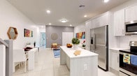 This contemporary two-story residence is defined by a clean, modern aesthetic characterized by crisp white walls, large-format tile flooring, and recessed lighting throughout. The main level showcases an open-concept layout where the kitchen, dining, and living areas flow seamlessly together. The kitchen is well-equipped with stainless steel appliances, including a French door refrigerator, microwave, range, and dishwasher, complemented by light wood-grain cabinetry extending to the ceiling and a spacious island with a white quartz countertop. Adjacent to the kitchen, a compact dining area features a white table and chairs, a hexagonal wood-framed mirror, and colorful wall art. The living area offers a low-profile media console and accent seating on large blue area rug. A staircase with white balusters and gray-toned treads connects to the upper level, highlighted by a skylight above. The bedrooms are bright and well-sized, each with tile flooring, roller shades, and vibrant artwork. One bedroom includes glass doors opening to a green outdoor view, while another features an en-suite bathroom. Bathrooms are finished with tile surrounds, tub-shower combinations, and wood-toned vanities with white countertops, maintaining the home's cohesive, contemporary style.