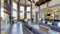 This impressive single-story space captivates from the moment you enter, defined by its dramatic vaulted ceilings adorned with dark, rustic exposed wood beams that create a bold architectural contrast against the crisp white ceiling panels. Towering multi-pane windows flood the interior with abundant natural light, framing views of the surrounding exterior and lending the space an open, airy feel.

The main gathering area is elegantly appointed with plush gray sofas arranged around a patterned area rug in cool teal and ivory tones, anchored by a reclaimed wood coffee table. A statement Sputnik-style chandelier hangs above, adding a modern, artistic touch that complements the space's blend of contemporary and rustic design elements.

A dedicated seating nook near the expansive windows offers a quieter setting with traditional wood furniture, ideal for individual use or small group gatherings. Rich, dark hardwood flooring flows throughout, grounding the space and enhancing its sophisticated ambiance.

To one side, a windowed interior room — partially visible through large sliding glass panels — extends the functionality of the space, hinting at additional private or semi-private areas. Neutral gray and taupe wall tones, paired with thoughtful accent lighting, complete this well-designed and welcoming environment.