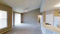 This single-floor residence offers a thoughtfully designed open-concept layout that maximizes both space and functionality. The main living area immediately draws the eye upward with its vaulted ceilings, which lend a sense of volume and airiness to the room. Soft, neutral-toned walls in warm beige complement the plush wall-to-wall carpeting underfoot, creating a comfortable and cohesive atmosphere throughout.

Natural light pours in generously through a large sliding glass door fitted with vertical blinds, providing easy access to an outdoor area while brightening the interior. A ceiling fan is centrally positioned to ensure year-round comfort. The open flow between the living space and the kitchen is a standout feature, with the kitchen area showcasing light stone countertops and a sink that overlooks the main living area — ideal for entertaining or everyday convenience.

A doorway visible from the main room leads to additional areas of the unit, suggesting a practical and well-connected floor plan. The overall design balances simplicity and warmth, making it a versatile and welcoming space suited for a range of lifestyles. Clean white trim throughout adds a crisp finishing touch to the neutral palette.