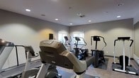 This fitness room offers a dedicated space for cardiovascular exercise, outfitted with multiple Matrix-brand machines, including elliptical trainers and a stair climber. The equipment is arranged to allow comfortable circulation throughout the room, with durable low-pile carpet flooring providing stable footing during workouts. Neutral gray walls create a clean, focused environment, while recessed lighting ensures consistent illumination throughout the space. Two windows along the far wall introduce natural light, contributing to an open and energizing atmosphere. A ceiling-mounted HVAC vent supports comfortable climate control during exercise sessions. The overall design is practical and functional, prioritizing equipment accessibility and ease of movement. The space is well-suited for cardio-focused training, with the Matrix equipment lineup reflecting a commitment to quality fitness amenities.