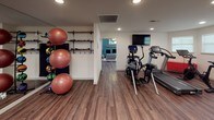 This single-level fitness space is thoughtfully designed to support a full range of training activities within a clean, well-organized layout. The flooring throughout is a warm hardwood-style laminate that provides both durability and an inviting aesthetic, tying the various workout zones together cohesively.

The cardio area is outfitted with a treadmill and a Cybex elliptical trainer, positioned beneath a wall-mounted flat-screen television for entertainment during workouts. Adjacent to this zone, a dedicated strength training area features a multi-station cable machine alongside an adjustable weight bench, offering versatile resistance training options. A large wall mirror expands the sense of space and allows for form checks during exercise.

An accessory wall displays a well-organized collection of stability balls, medicine balls, and resistance tools on wooden shelving, keeping the floor area open and functional. Recessed ceiling lighting illuminates the space evenly, and a skylight in one area introduces natural light. The walls feature a lively patterned wallcovering in the main training zone, adding visual personality. Multiple entry points with glass-paneled doors allow for natural light and easy access throughout the space.
