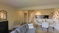 This single-floor unit offers an open and cohesive layout that seamlessly connects the living, dining, and kitchen areas. The living room serves as the central gathering space, featuring light-toned carpeting, neutral cream walls, and enough room for a full sofa arrangement alongside accent seating. A patterned armchair adds a decorative touch near the entry side of the room.

Toward the rear of the living area, a compact dining nook accommodates a small table and chairs, positioned adjacent to a kitchen counter with bar-height seating. The kitchen is distinguished by a striking mosaic tile backsplash in blue and metallic tones, which provides a notable design accent against the otherwise neutral palette. The counter pass-through enhances the open feel between the kitchen and living spaces.

A hallway visible from the main room leads to additional areas of the unit, with a closet featuring a rod and hangers visible through an open door, suggesting convenient storage. A secondary door with a slatted panel design adds architectural interest to the corridor. Overhead lighting illuminates the hallway, contributing to a bright and navigable interior throughout the space.