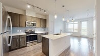 Located at 2810 Babcock Rd in San Antonio, TX, this one-bedroom, one-bathroom unit offers a well-designed, open-concept layout that maximizes every square foot of living space. The kitchen is a standout feature, showcasing rich dark cabinetry, stainless steel appliances including a refrigerator, range, and built-in microwave, and a striking central island with a white countertop and undermount sink. Elegant pendant lighting hangs above the island, complementing the track lighting overhead. Wood-style flooring flows throughout the main living and kitchen areas, creating a warm and cohesive aesthetic. The living area benefits from large windows and a ceiling fan, filling the space with natural light and views of the surrounding greenery. A private balcony extends the living space outdoors, offering scenic, tree-lined views of the San Antonio skyline corridor. The bedroom is carpeted for comfort, fitted with a ceiling fan, and receives ample natural light through a well-placed window. The bathroom features wood-style tile flooring, a dark vanity with granite countertops, dual mirrors with overhead lighting, and a tub-shower combination with clean white subway tile surround. This unit combines modern finishes with functional design throughout.