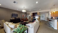 Located at 4175 Wagner St in Eugene, Oregon, this thoughtfully designed single-floor community space offers a warm and inviting atmosphere throughout its various functional areas. The main lounge is the centerpiece of the space, anchored by a striking floor-to-ceiling stone and tile fireplace with a dark wood mantel, creating a cozy focal point. Multiple seating arrangements are thoughtfully positioned throughout, including plush cream sofas with accent pillows, rich cognac leather armchairs, and accent chairs — all set atop neutral carpeting that extends across the space. A wall-mounted flat-screen television and a sleek media console add a media-ready component to the lounge area. Recessed lighting and pendant fixtures provide consistent, comfortable illumination throughout. A secondary seating area near the entrance features large windows with wood-slatted blinds, allowing natural light to filter in through double glass doors. The kitchen is well-equipped with warm wood cabinetry, dark granite countertops, a mosaic tile backsplash, stainless steel appliances including a refrigerator, range, and built-in microwave, and tile flooring. Adjacent to the kitchen, a dedicated dining area accommodates round tables with dark leather chairs, ideal for group gatherings. The overall ambiance is modern and comfortable, with a cohesive earthy color palette unifying the entire space.