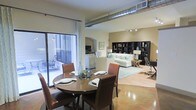 This single-floor space showcases a compelling blend of industrial and contemporary design. Exposed metallic ductwork runs across the ceiling, lending an urban loft aesthetic that is complemented by warm, neutral-toned walls and polished concrete floors that reflect light throughout the open layout.

The dining area anchors one end of the space, centered around a round wooden table with rich brown leather chairs, set atop the gleaming floor. A large sliding glass door floods this area with natural light and provides direct access to an outdoor patio furnished with seating, seamlessly connecting interior and exterior living.

The open floor plan flows naturally into the living area, where a light-colored sectional sofa is arranged on a soft area rug, creating a comfortable and inviting lounge space. Track lighting above highlights the room's features, while an arched architectural detail adds a classic touch that contrasts elegantly with the industrial elements overhead.

A built-in bookshelf unit along the far wall offers both display and storage space, styled with decorative objects and books. A dedicated workspace is also visible, making this space as functional as it is visually appealing. The kitchen area is partially visible, integrated smoothly into the open-concept design. Overall, the space balances practicality with a refined, modern sensibility.