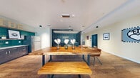 Located at 1100 D St in Las Vegas, NV, this thoughtfully designed single-floor communal space offers a well-appointed and versatile layout. The open floor plan is anchored by a stunning live-edge wood dining table with leather chairs, serving as a natural centerpiece beneath sleek pendant lighting. Warm hardwood flooring flows throughout, tying the space together with an inviting, cohesive aesthetic.

A floor-to-ceiling glass wall system with sliding doors floods the interior with abundant natural light and provides seamless access to an outdoor patio area, extending the usable space and enhancing the connection between indoors and out. Adjacent to the dining area is a compact kitchenette finished with teal tile backsplash and dark cabinetry, adding a vibrant design accent.

A vertical slat room divider elegantly separates the dining and lounge zones without fully enclosing them, maintaining an open and airy feel. The lounge area features a generous sectional sofa in cool blue-grey tones, complemented by a plush area rug and muted blue accent walls adorned with curated artwork. Built-in cabinetry and a window bench offer additional functionality. The overall ambiance is modern and welcoming, making this space well-suited for gathering, collaboration, or relaxation.