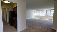 This single-floor residence offers a straightforward and functional layout designed for comfortable everyday living. The open living area is finished with wall-to-wall carpet flooring that extends throughout, creating a warm and cohesive feel underfoot. A large window dressed with vertical blinds dominates one wall of the living space, allowing an abundance of natural light to fill the room and lending the area a bright, airy atmosphere. Neutral-toned walls and clean white baseboards keep the aesthetic simple and versatile.

Adjacent to the living area, the kitchen is partially separated by a structural partition wall, providing a sense of definition between the two spaces while maintaining an open connection. The kitchen features dark wood cabinetry with brushed metal hardware, offering ample storage for cookware and pantry essentials. The countertops are finished in a dark stone or stone-look surface, adding a touch of contrast against the cabinetry. Tile flooring in the kitchen area provides a practical and easy-to-clean transition from the carpeted living space.

Overall, the residence presents a clean, move-in-ready environment with classic finishes and a practical single-level floor plan that balances openness with defined functional zones.