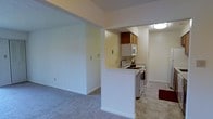 This single-floor space showcases a practical, open-concept design that connects the living area and kitchen in a seamless layout. The living area features soft, neutral-toned carpeting that adds warmth underfoot, while clean white walls contribute to a bright and airy atmosphere throughout.

The kitchen is positioned behind a half-wall partition, creating a natural visual separation while maintaining an open feel. It is equipped with white appliances, including an electric range, overhead microwave, and additional upper cabinetry in a warm wood finish that provides ample storage. The kitchen floor transitions to a hard surface material, offering a practical contrast to the carpeted living space.

Flush ceiling lighting ensures consistent illumination across the kitchen and adjoining areas. The half-wall peninsula serves both as a functional boundary and an informal breakfast bar-style surface. Neutral paint tones are carried throughout, reinforcing a cohesive and understated aesthetic. The overall layout is efficiently designed, making effective use of available space while maintaining a clean and well-organized appearance.