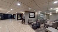 Located on S Chatham Ave in Elmhurst, Illinois, this single-floor lobby space presents a polished and welcoming commercial interior designed with a contemporary aesthetic. The space is finished with large-format cream tile flooring throughout, providing a clean and cohesive foundation that reflects the recessed lighting above.

The main gathering area features a well-appointed seating arrangement with upholstered gray armchairs and accent ottomans arranged over a patterned area rug, creating an inviting lounge atmosphere. Framed landscape artwork adorns the taupe-toned walls, complemented by elegant wall sconces that cast a warm ambient glow. A full-length mirror with a bold dark wood frame and a decorative console table with sculptural branch arrangements add refined touches to the corridor areas.

The lobby corridor is notably spacious, with a dropped acoustic tile ceiling fitted with recessed lighting that illuminates the open walkway. A standout feature is the elevator vestibule, clad in dramatic dark stacked-stone tile surrounding a sleek black elevator door — a striking architectural contrast to the otherwise neutral palette. The hallway beyond leads to additional rooms, maintaining the same consistent, professional design language throughout. The overall ambiance is modern, refined, and functional, well-suited for a commercial or multi-use professional setting.