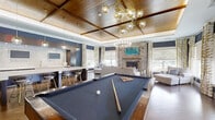 This 750 square foot single-floor space seamlessly blends modern luxury with cozy comfort. The heart of the area is a stunning living room, anchored by a floor-to-ceiling stone fireplace that adds rustic charm and warmth. Large windows with geometric patterned curtains flood the space with natural light, while offering views of the surrounding area.

The living room features a plush sectional sofa and chaise lounge, perfectly positioned on a vibrant blue area rug. A vaulted ceiling with exposed wooden beams adds height and character, complemented by a striking circular chandelier.

Adjacent to the living area is a state-of-the-art kitchen. It boasts a large island with sleek white countertops and modern bar stools, ideal for casual dining or entertaining. Pendant globe lights hang above, providing both style and illumination. The kitchen's dark cabinetry contrasts beautifully with the light countertops and backsplash, creating a sophisticated aesthetic.

The space's cohesive design is evident in the consistent use of materials and colors throughout, from the hardwood floors to the navy blue accent walls. This thoughtfully designed area maximizes its 750 square feet, offering a perfect blend of functionality and style for modern living.