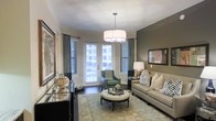 This single-floor space showcases a thoughtfully designed living area that blends comfort with refined style. The living room serves as the centerpiece, featuring dark hardwood flooring that anchors the space and contrasts beautifully with the crisp white ceiling and warm olive-toned accent wall. A statement drum pendant light hangs centrally, casting a warm glow over the seating arrangement below. Natural light floods the room through multiple windows fitted with horizontal blinds and floor-length drapes, while a set of glass-paned French doors opens to an outdoor area, enhancing the sense of openness. The room is furnished with a neutral-toned sofa accented by geometric throw pillows, a tufted round ottoman serving as a central coffee table, and a complementary accent chair in muted green. A floral-patterned area rug defines the seating zone, adding texture and color to the space. Wall art and a sideboard with metallic accents contribute to the polished, curated aesthetic. The overall ambiance is inviting and sophisticated, with a harmonious balance of natural light, warm tones, and contemporary furnishings that make the space feel both functional and elegantly appointed.