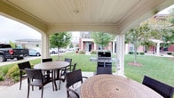 This single-level outdoor space offers a thoughtfully designed covered patio that serves as a communal gathering and entertaining area. The structure features a classic beadboard ceiling supported by painted columns, providing shade and shelter while maintaining an open, airy feel. The patio accommodates multiple round dining tables paired with wicker-style chairs, creating comfortable seating arrangements ideal for outdoor meals and socializing. A full-sized gas grill is conveniently positioned within the covered area, making outdoor cooking accessible and practical. The space opens directly onto a well-kept lawn framed by mature trees, lending a peaceful, park-like atmosphere. Beyond the greenery, a brick residential building is visible, reflecting the broader community setting. The combination of durable outdoor furnishings, functional grilling equipment, and lush landscaping makes this patio a versatile space suited for casual dining, social gatherings, and everyday outdoor relaxation. The covered design ensures the area remains usable in a variety of weather conditions, enhancing its appeal as a year-round amenity within the community.