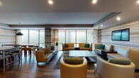 This single-floor communal space is thoughtfully designed with a contemporary aesthetic, combining comfort and functionality across several distinct zones. The lounge area is the centerpiece, featuring an arrangement of tan leather sofas and chairs accented with rich teal throw pillows, all anchored by a multi-tiered rectangular coffee table with a warm wood-tone finish. Floor-to-ceiling windows line the exterior walls, flooding the space with natural light and offering views of surrounding greenery.

A signature design element throughout is the horizontally striped wall treatment in warm tones of beige, tan, and gray, lending visual texture and cohesion across zones. Geometric pendant light fixtures hang above the seating area, adding a sculptural focal point alongside a wall-mounted flat-screen television.

The space transitions into a casual dining and bar area with high-top seating and a kitchen nook outfitted with sage-green cabinetry, stainless steel appliances including a full-size refrigerator and microwave, and white countertops. Curated artwork, including a bold botanical painting, adds personality to the walls. Hardwood-style flooring and recessed lighting complete the polished, welcoming ambiance throughout.